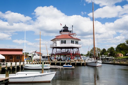 A lighthouse on stilts by the water, surrounded by boats and a blue sky with fluffy clouds.