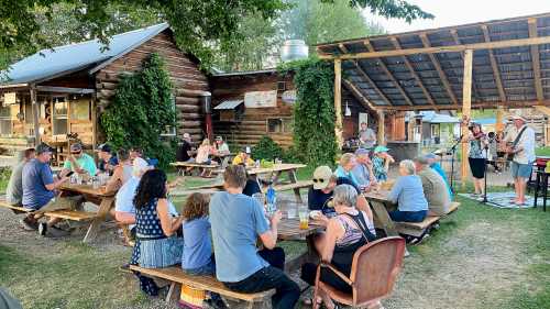 A lively outdoor gathering at a rustic venue, with people seated at picnic tables enjoying food and drinks while a musician performs.