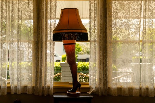 A lamp shaped like a woman's leg with fishnet stockings, set against lace curtains and a sunny outdoor view.