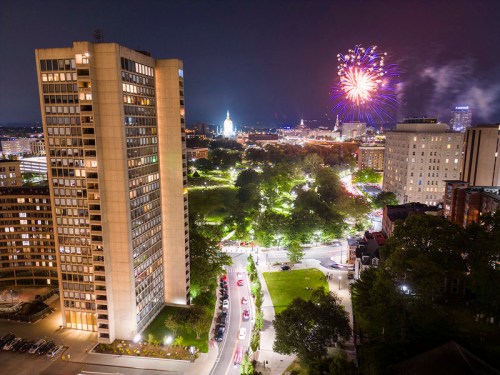 A nighttime cityscape featuring fireworks over a park, with illuminated buildings and streets below.