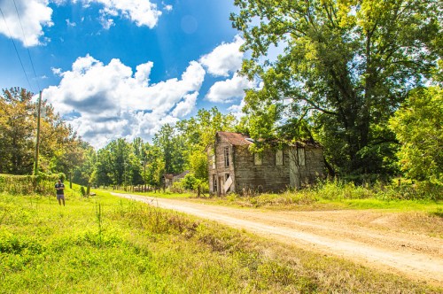 A dirt road leads past an abandoned, weathered building surrounded by lush greenery and a bright blue sky.