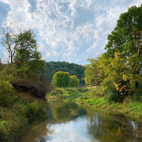 A serene river flows through lush greenery under a cloudy sky, surrounded by trees and rolling hills.