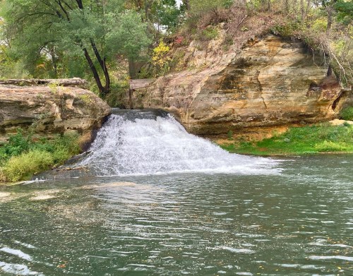 A small waterfall cascades over rocks into a serene green pool, surrounded by trees and lush vegetation.