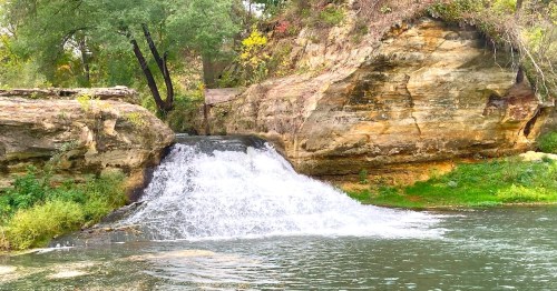 A serene waterfall cascading over rocks into a calm river, surrounded by lush greenery and trees.