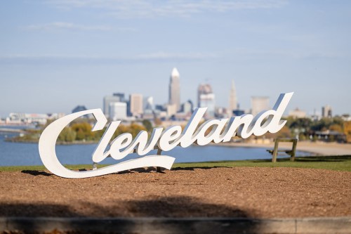 A large, white "Cleveland" sign in the foreground with the city skyline and lake in the background on a clear day.