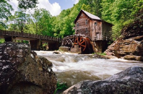 A historic wooden mill with a water wheel beside a rushing river, surrounded by lush green trees and rocky terrain.