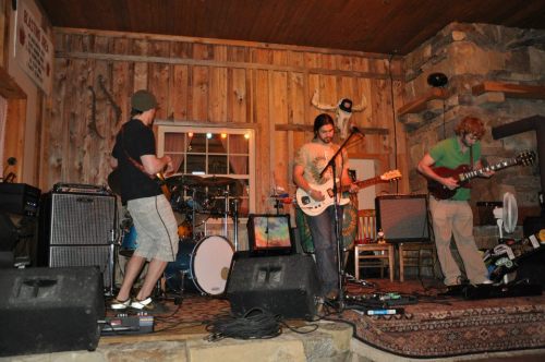 A band performs on stage in a rustic venue with wooden walls and instruments, creating a lively atmosphere.