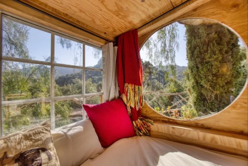Cozy interior of a tiny home with a round window, colorful pillows, and a scenic view of greenery outside.