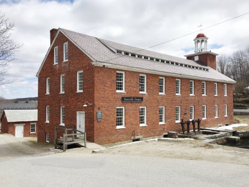 Historic brick building with a bell tower, surrounded by trees and a gravel path, under a cloudy sky.