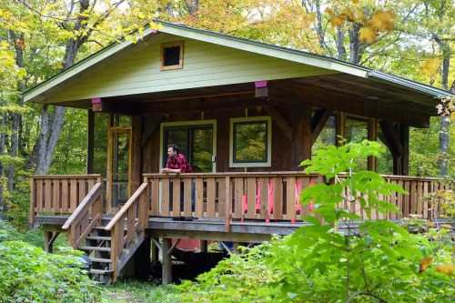 A cozy wooden cabin surrounded by trees, with a person in a red flannel shirt on the porch.