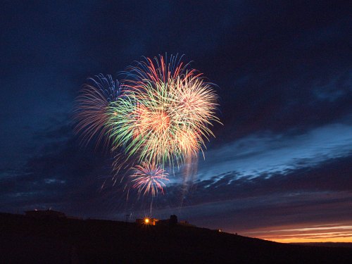 Colorful fireworks burst in the night sky, illuminating clouds against a dark blue backdrop at sunset.