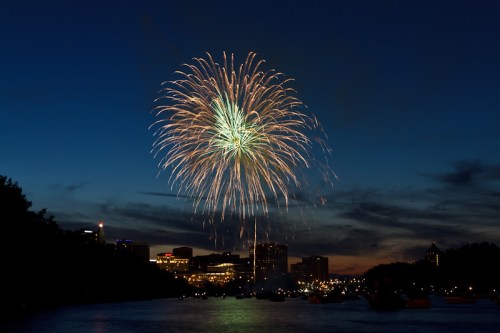 Colorful fireworks burst over a city skyline at dusk, reflecting on the water below.