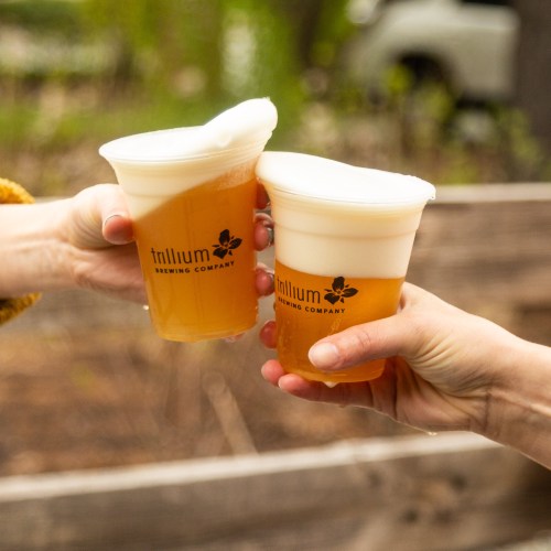 Two hands clink plastic cups of beer, featuring the Trillium Brewing Company logo, against a blurred outdoor background.