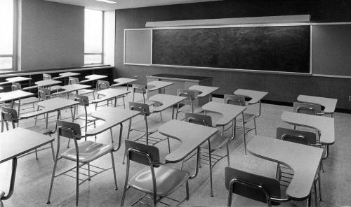 A black and white image of an empty classroom with desks and chairs arranged facing a chalkboard.