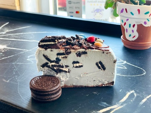 A slice of ice cream cake topped with crushed cookies and colorful candies, next to a stack of Oreo cookies.