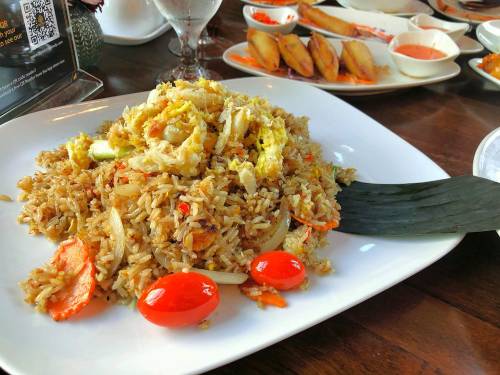 A plate of fried rice with vegetables, shrimp, and cherry tomatoes, served on a white dish with a black spatula.