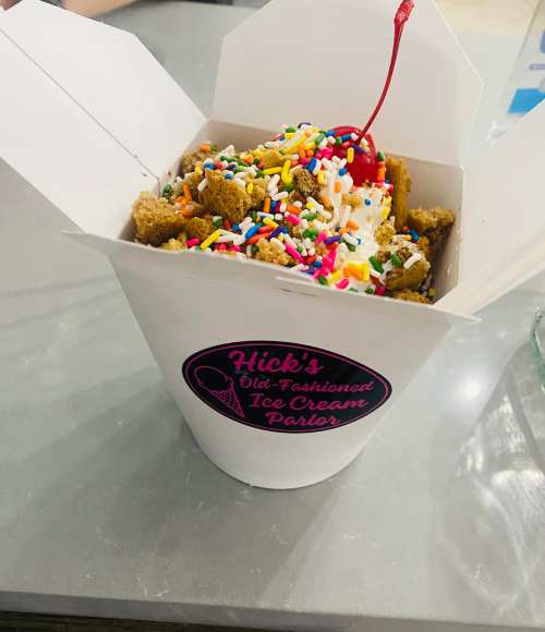 A takeout box filled with ice cream, whipped cream, sprinkles, and a cherry on top, sitting on a gray table.