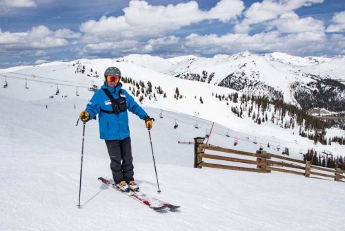 A skier in a blue jacket stands on a snowy slope with mountains and ski lifts in the background under a cloudy sky.