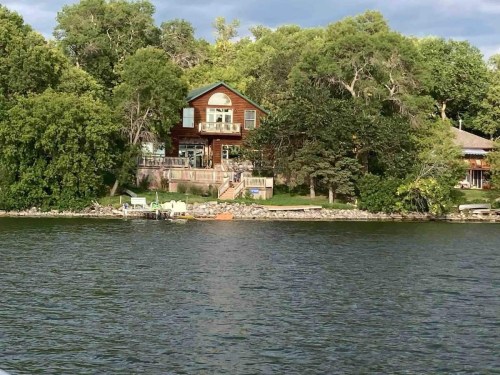 A lakeside house surrounded by trees, with a deck and a small dock visible along the water's edge.