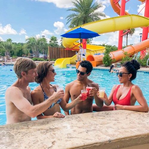 Four friends enjoy drinks by a pool with a water slide in the background, surrounded by palm trees and sunshine.