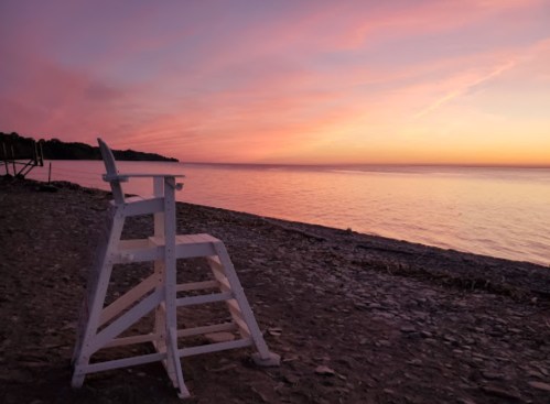 A white lifeguard chair on a beach at sunset, with colorful skies reflecting on calm water.