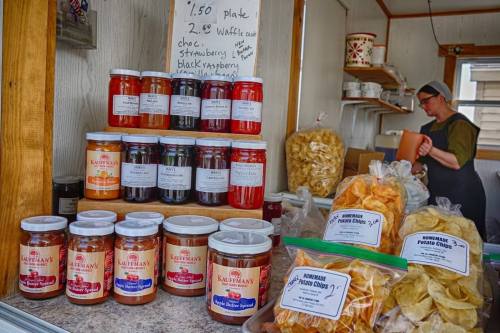A display of jars filled with jams and bags of snacks at a market stall, with a vendor in the background.