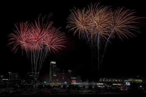 Colorful fireworks burst in the night sky above a city skyline, illuminating the dark with vibrant reds and golds.