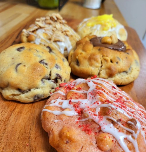 A variety of large cookies on a wooden surface, including chocolate chip, cinnamon, lemon, and strawberry flavors.
