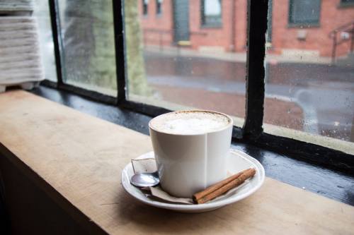A cup of coffee with cinnamon sticks sits on a windowsill, with a rainy street view outside.
