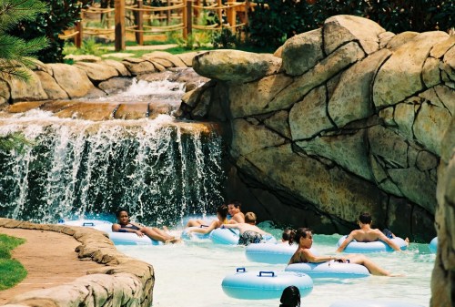 People float on inner tubes in a lazy river, surrounded by rocks and a waterfall in a sunny outdoor setting.