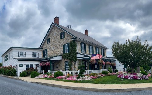 A charming stone building with green shutters, surrounded by colorful flowers and an American flag, under a cloudy sky.