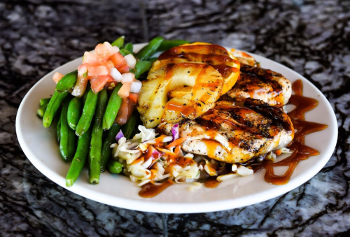 Grilled chicken with pineapple, green beans, and coleslaw drizzled with barbecue sauce on a white plate.