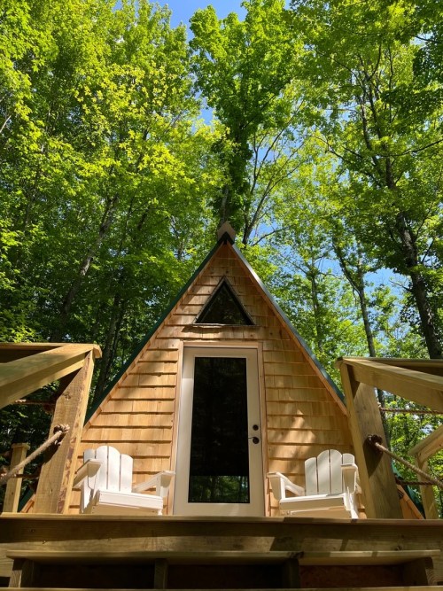 A cozy A-frame cabin nestled in a lush green forest, with two white chairs on the porch.