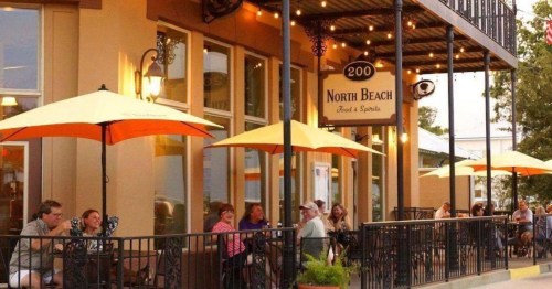Outdoor dining scene at North Beach with patrons under yellow umbrellas, enjoying food and drinks on a sunny day.