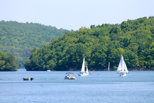 Sailboats and motorboats on a calm lake surrounded by lush green hills under a clear blue sky.