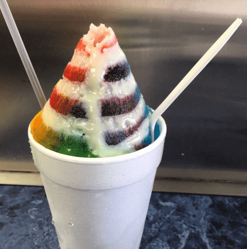 A colorful rainbow snow cone in a white cup, topped with a drizzle of syrup and two plastic spoons.