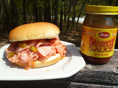 A sandwich with ham and barbecue sauce on a plate, next to a jar of barbecue sauce, set in a natural outdoor setting.