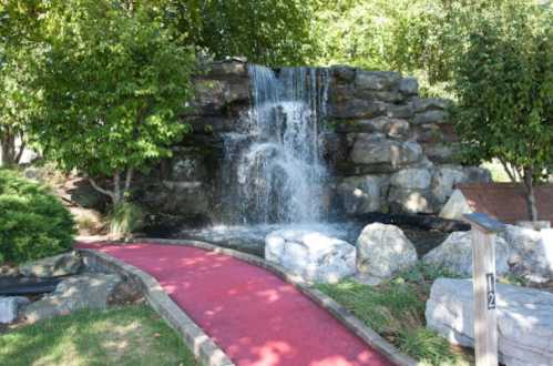 A small waterfall cascades over rocks, with a red pathway leading through lush greenery.