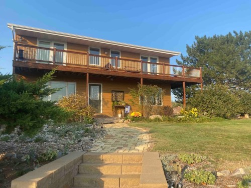 A two-story wooden house with a balcony, surrounded by greenery and a stone pathway leading to the entrance.