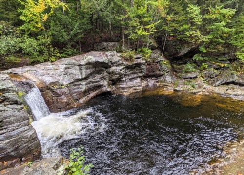 A serene waterfall cascades into a dark pool, surrounded by rocky terrain and lush green trees.