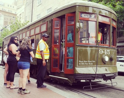 A vintage streetcar with passengers boarding, set in an urban environment. A conductor assists at the entrance.