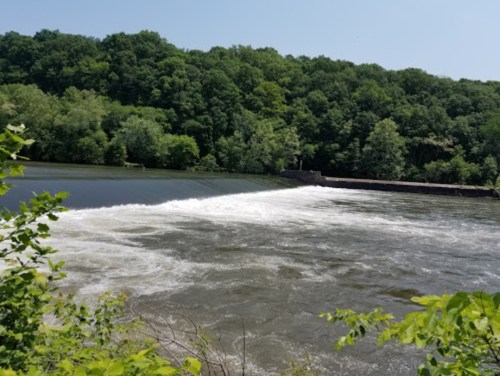 A serene river scene with a dam, flowing water, and lush green trees in the background under a clear blue sky.