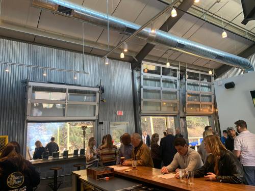 A lively brewery interior with large windows, metal walls, and patrons enjoying drinks at a long wooden bar.