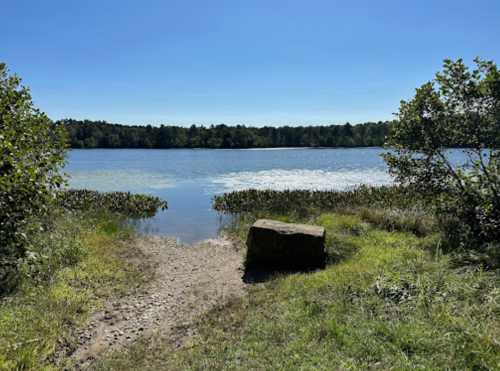 A serene lake view with a clear blue sky, surrounded by greenery and a large rock in the foreground.