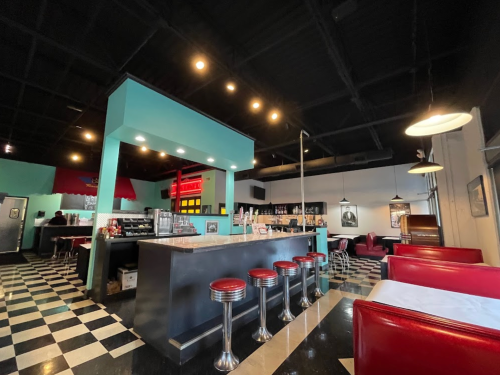 A retro diner interior featuring a checkered floor, bar stools, and colorful decor with a counter and seating area.