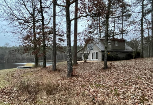 A cozy house by a pond, surrounded by trees and fallen leaves, on a cloudy day.