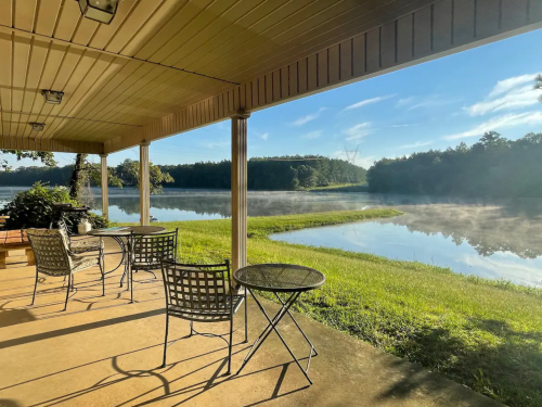 A serene lakeside view from a porch, featuring chairs and a table, with mist rising over the water in the morning light.