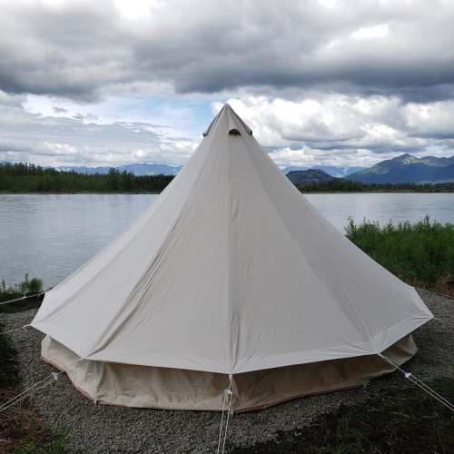 A large white tent by a river, surrounded by greenery and mountains under a cloudy sky.