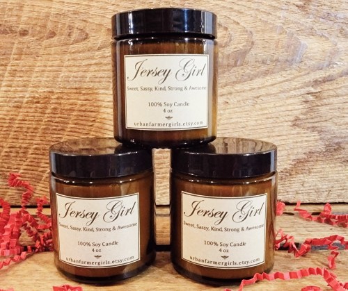 Three brown jars of "Jersey Girl" soy candles stacked on a wooden surface, labeled with sweet and sassy descriptions.