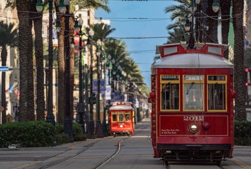 Two red streetcars on a palm-lined street, with one approaching and the other in the distance. Bright blue sky above.